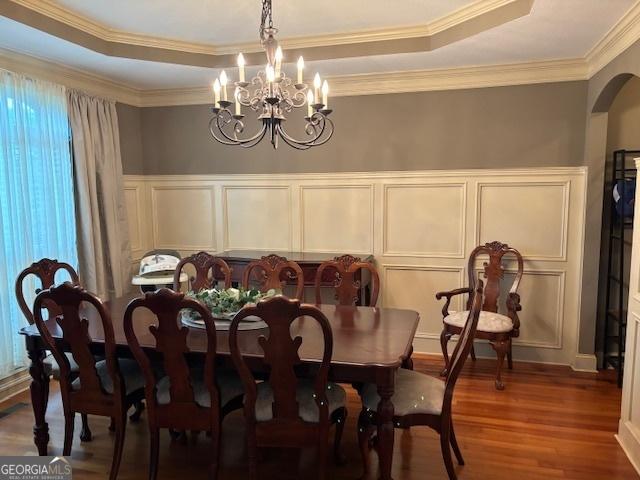 dining area featuring a decorative wall, a raised ceiling, wood finished floors, a chandelier, and wainscoting