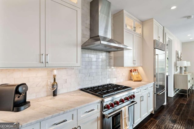 kitchen with high quality appliances, wall chimney exhaust hood, light stone countertops, dark wood-style flooring, and glass insert cabinets