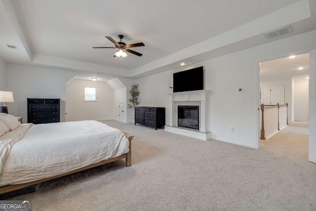bedroom featuring baseboards, a tray ceiling, light carpet, a glass covered fireplace, and a ceiling fan