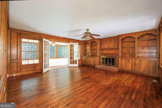 unfurnished living room featuring dark wood-type flooring, built in shelves, a glass covered fireplace, ceiling fan, and french doors