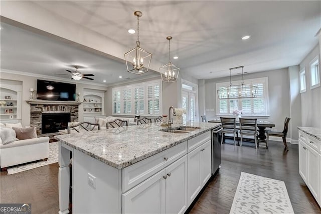 kitchen with a sink, stainless steel dishwasher, a fireplace, a center island with sink, and light stone countertops