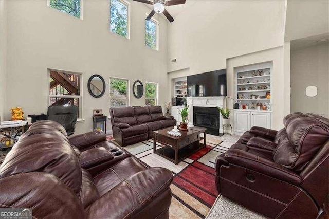 living room featuring a ceiling fan, built in shelves, a towering ceiling, and a fireplace with flush hearth