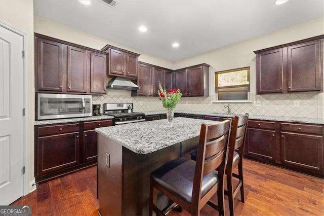 kitchen featuring appliances with stainless steel finishes, under cabinet range hood, a kitchen bar, dark wood-style flooring, and a kitchen island