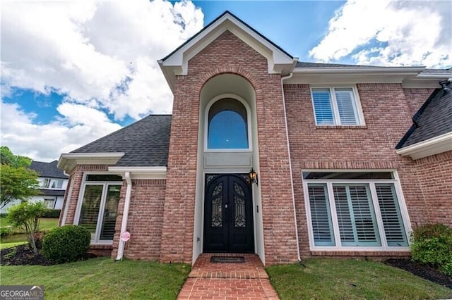 entrance to property featuring french doors, brick siding, a yard, and a shingled roof