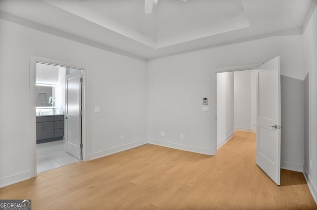unfurnished bedroom with light wood finished floors, a tray ceiling, baseboards, and connected bathroom