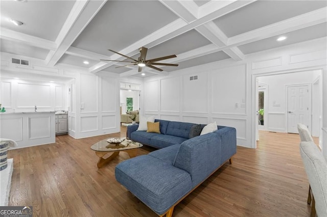 living area with a decorative wall, coffered ceiling, a ceiling fan, beam ceiling, and light wood-style flooring
