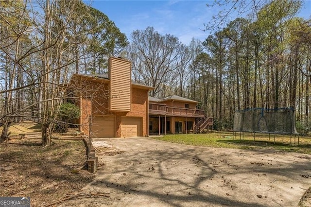 rear view of property with a trampoline, concrete driveway, a wooden deck, an attached garage, and a chimney