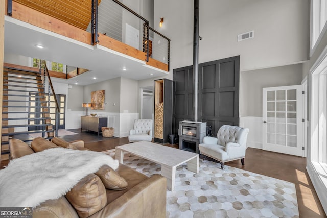 living room featuring a towering ceiling, stairway, a decorative wall, a wood stove, and a wainscoted wall