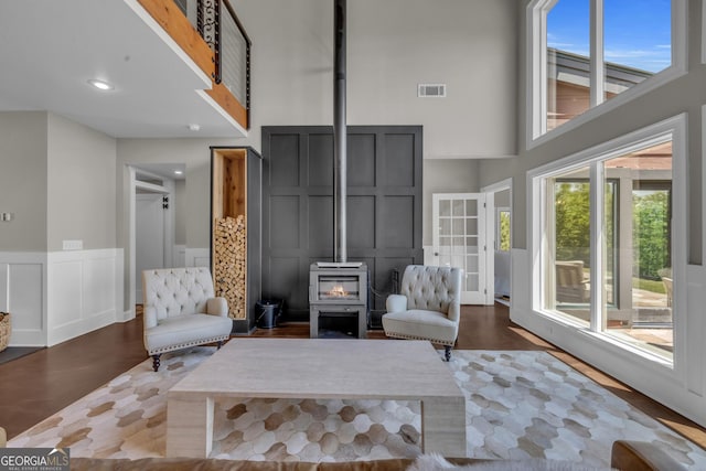 living room featuring a high ceiling, a wood stove, a decorative wall, wood finished floors, and a wainscoted wall