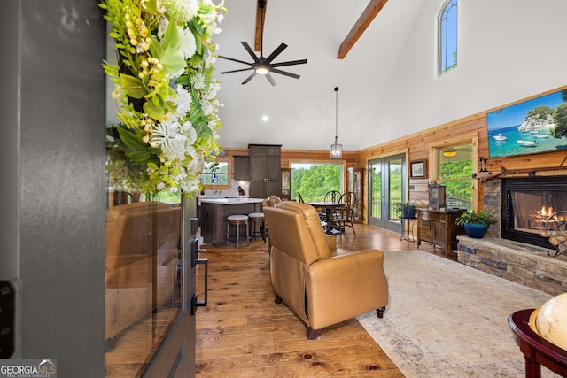 living room featuring high vaulted ceiling, ceiling fan, light wood finished floors, a stone fireplace, and beamed ceiling