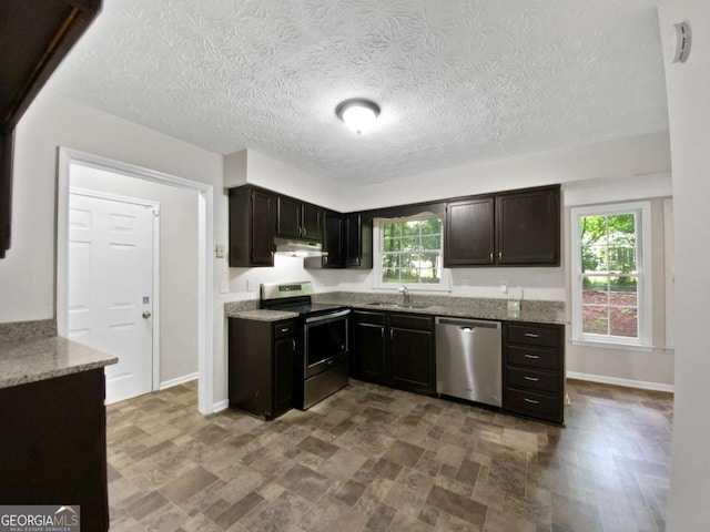 kitchen featuring appliances with stainless steel finishes, a sink, light stone countertops, under cabinet range hood, and baseboards