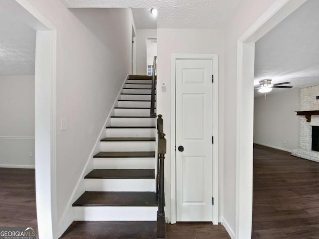 stairs with ceiling fan, wood finished floors, a large fireplace, baseboards, and a textured ceiling
