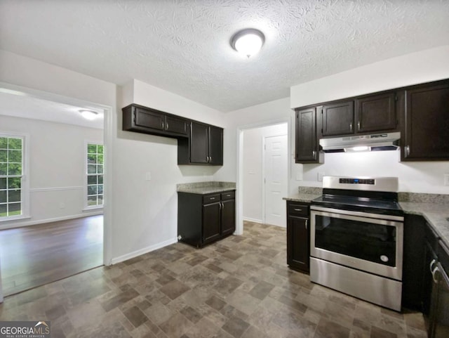 kitchen with stainless steel range with electric cooktop, under cabinet range hood, baseboards, stone finish floors, and dark brown cabinetry
