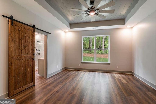 spare room featuring a barn door, a tray ceiling, wood finished floors, baseboards, and a ceiling fan