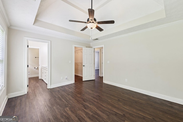 unfurnished bedroom with a tray ceiling, dark wood-type flooring, a walk in closet, baseboards, and crown molding