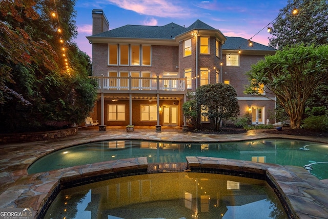 rear view of house featuring a chimney, a pool with connected hot tub, a patio, a balcony, and brick siding