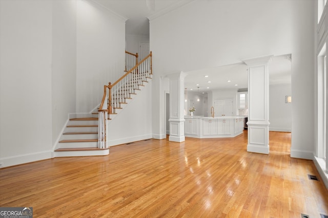 unfurnished living room featuring ornate columns, light wood finished floors, stairs, a towering ceiling, and crown molding