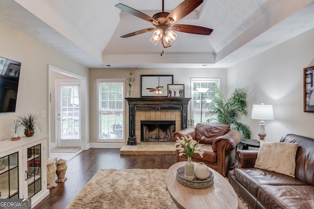 living area with a tray ceiling, a fireplace, a ceiling fan, dark wood-style flooring, and baseboards