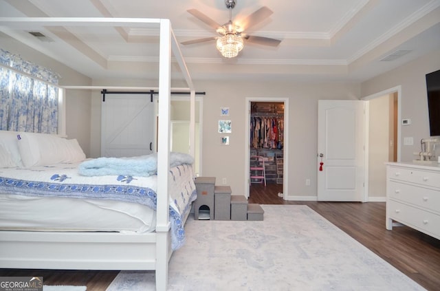 bedroom with a tray ceiling, wood finished floors, a walk in closet, crown molding, and a barn door