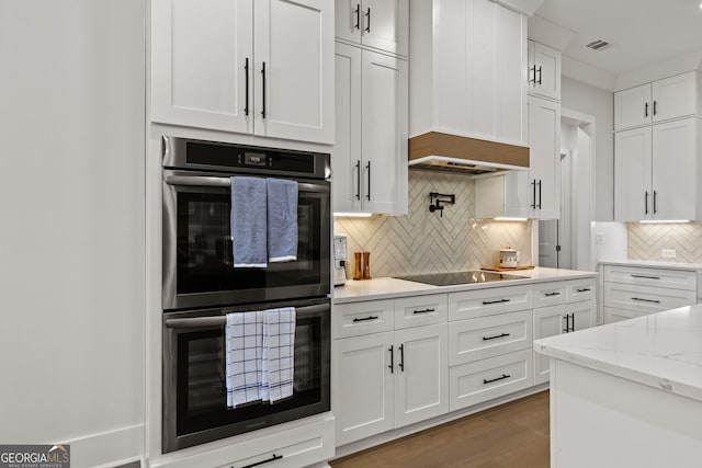 kitchen featuring stainless steel double oven, black electric cooktop, white cabinets, tasteful backsplash, and light stone countertops