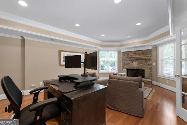 home office with light wood finished floors, a raised ceiling, crown molding, a stone fireplace, and recessed lighting