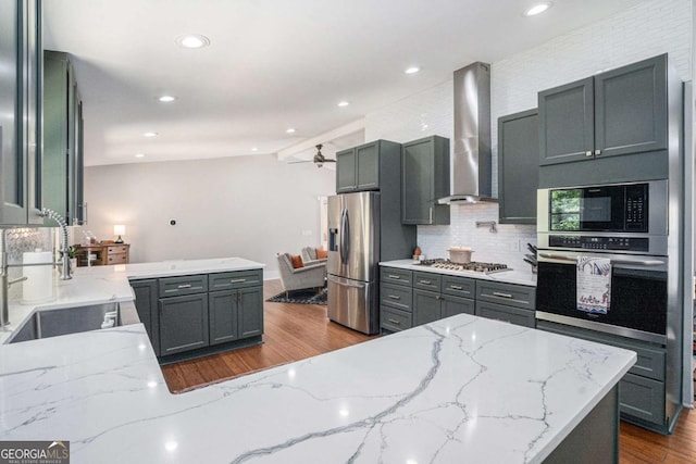 kitchen featuring appliances with stainless steel finishes, wall chimney exhaust hood, a sink, dark wood-style floors, and a peninsula
