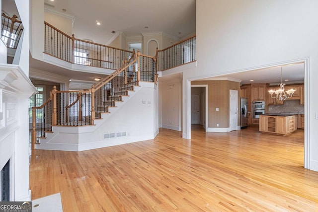 unfurnished living room featuring a towering ceiling, a chandelier, stairs, light wood-style flooring, and a fireplace with flush hearth