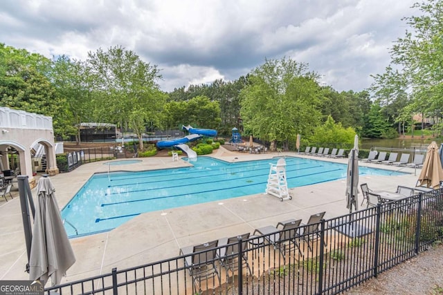 community pool featuring a patio area, fence, a water slide, and view of scattered trees