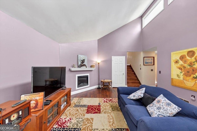 living area featuring high vaulted ceiling, stairway, wood finished floors, baseboards, and a glass covered fireplace