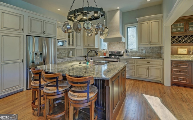 kitchen with stainless steel appliances, premium range hood, cream cabinets, a sink, and light stone countertops