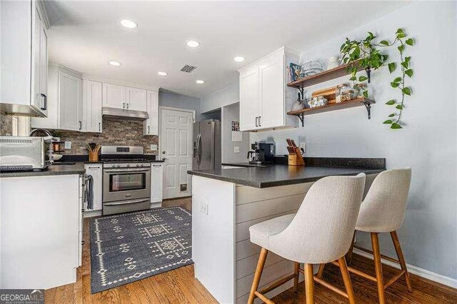 kitchen with appliances with stainless steel finishes, a peninsula, under cabinet range hood, white cabinets, and dark countertops