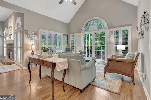 living area with high vaulted ceiling, ceiling fan, a fireplace with raised hearth, light wood-type flooring, and baseboards