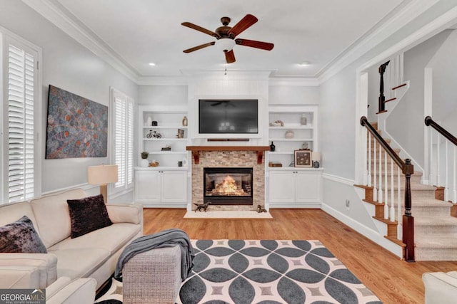 living room featuring light wood-style floors, a glass covered fireplace, ornamental molding, a ceiling fan, and built in features