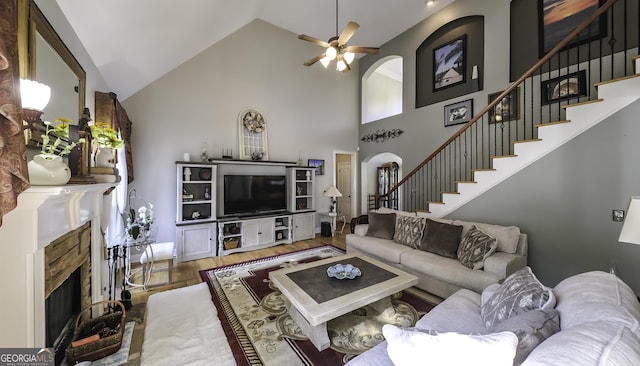 living room with a fireplace with raised hearth, stairway, ceiling fan, wood finished floors, and high vaulted ceiling