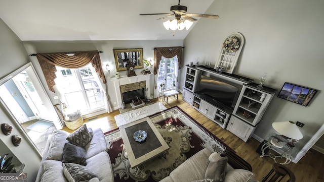 living area with wood finished floors, a stone fireplace, a ceiling fan, healthy amount of natural light, and lofted ceiling