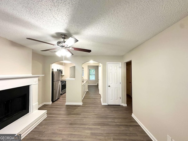 unfurnished living room featuring ceiling fan, a fireplace with raised hearth, dark wood finished floors, baseboards, and a textured ceiling