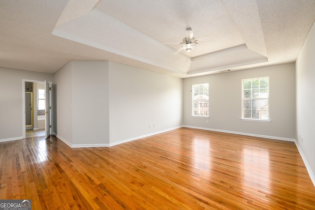 empty room with ceiling fan, a tray ceiling, light wood finished floors, a textured ceiling, and baseboards