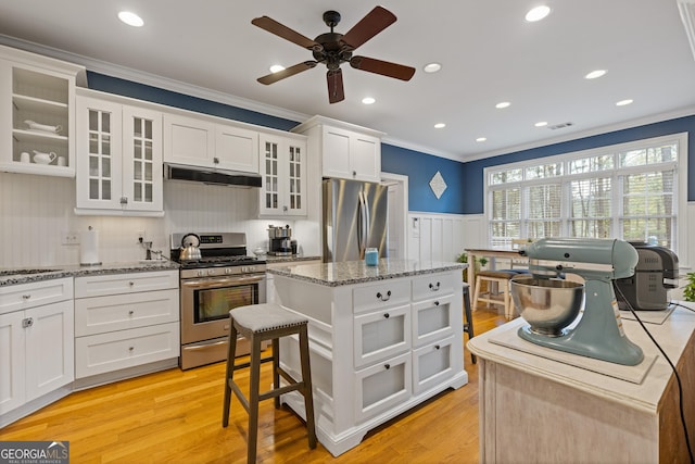 kitchen with stainless steel appliances, under cabinet range hood, ornamental molding, a wainscoted wall, and a ceiling fan