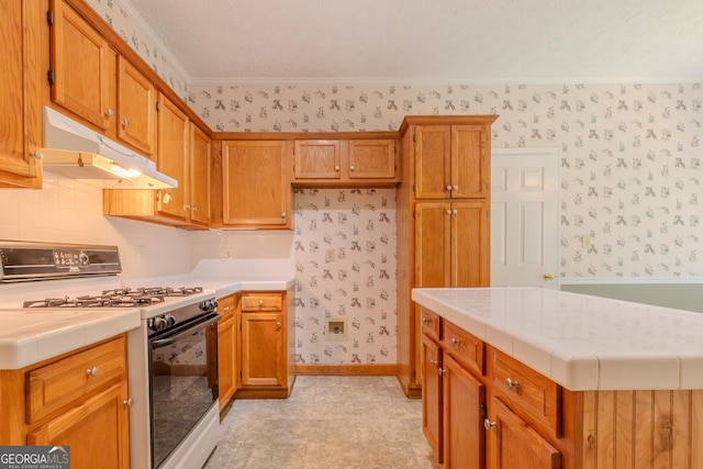 kitchen with range with gas cooktop, wallpapered walls, tile counters, under cabinet range hood, and brown cabinets