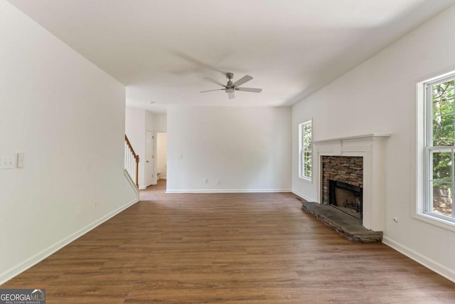 unfurnished living room featuring wood finished floors, a stone fireplace, ceiling fan, stairs, and baseboards