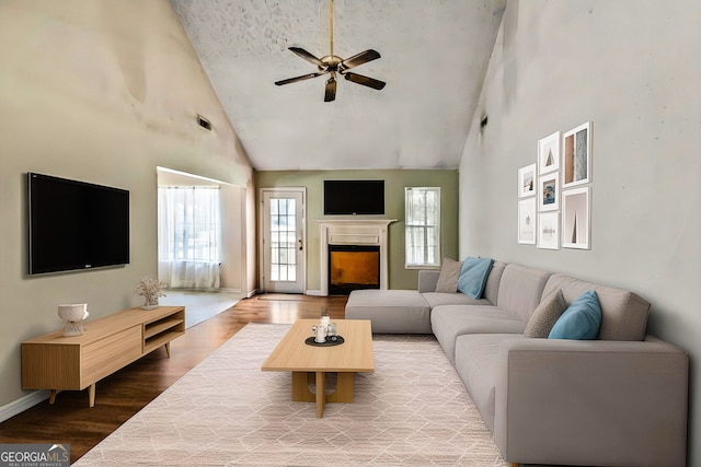 living area with high vaulted ceiling, wood finished floors, ceiling fan, a fireplace, and baseboards