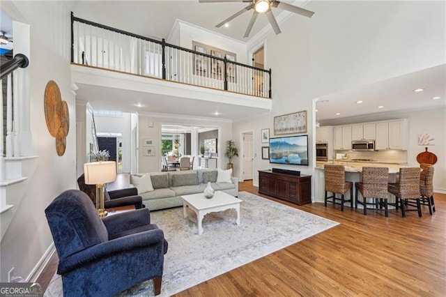 living room featuring ceiling fan, light wood-style floors, a towering ceiling, ornamental molding, and baseboards