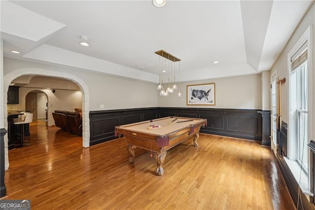 recreation room featuring a tray ceiling, pool table, light wood-style floors, arched walkways, and a decorative wall