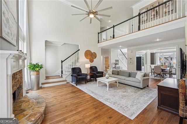 living room with stairway, a stone fireplace, a high ceiling, wood finished floors, and a ceiling fan