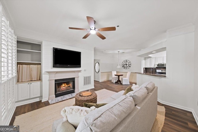 living room with a ceiling fan, dark wood-style floors, a glass covered fireplace, baseboards, and ornamental molding