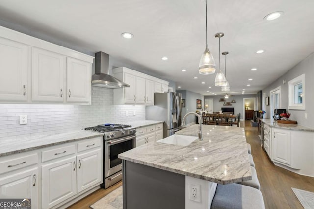 kitchen featuring wall chimney range hood, appliances with stainless steel finishes, a sink, open floor plan, and white cabinets