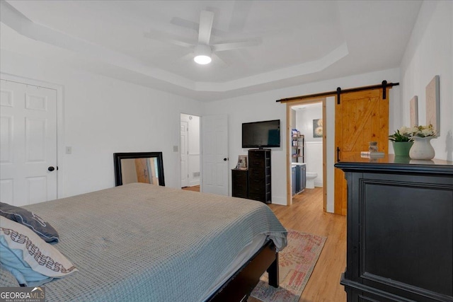 bedroom featuring a tray ceiling, a barn door, light wood finished floors, ensuite bath, and a ceiling fan