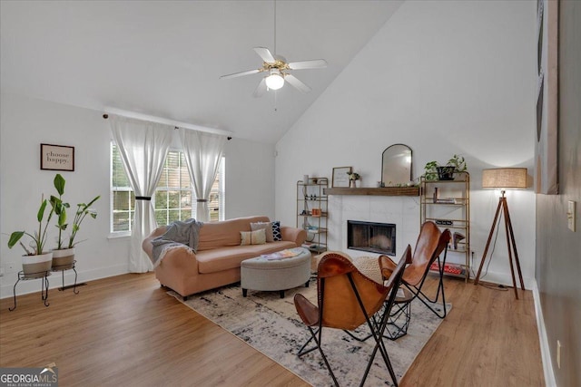 living room with ceiling fan, light wood-style flooring, high vaulted ceiling, a tiled fireplace, and baseboards