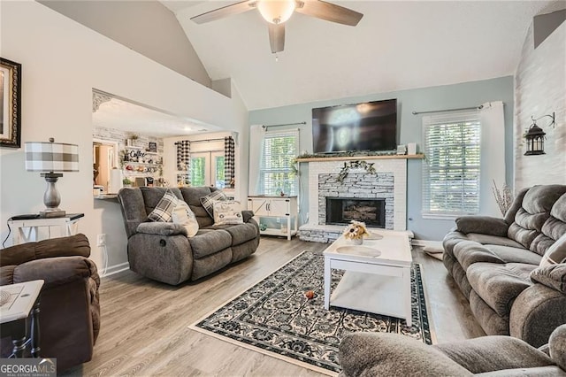 living area with a ceiling fan, wood finished floors, baseboards, a stone fireplace, and high vaulted ceiling