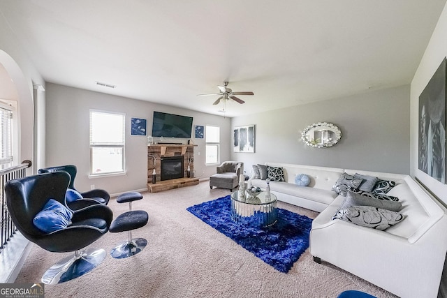 carpeted living room featuring a stone fireplace, a ceiling fan, baseboards, and arched walkways